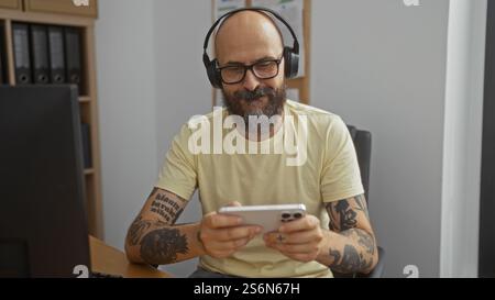 Homme hispanique barbu chauve avec des tatouages à l'aide d'un smartphone et portant des écouteurs dans un bureau Banque D'Images