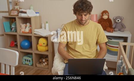 Jeune homme à l'aide d'un ordinateur portable dans une chambre confortable remplie de jouets pour bébé et d'un berceau, créant une atmosphère calme à l'intérieur de la maison, mettant en valeur un ADU attrayant Banque D'Images