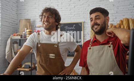 Deux boulangers masculins debout ensemble dans une boulangerie confortable, portant des tabliers et souriant, avec du pain fraîchement cuit en arrière-plan. Banque D'Images