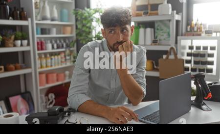 Jeune homme assis à un bureau dans un magasin de décoration intérieure avec diverses décorations et plantes à l'arrière-plan, regardant réfléchi et pensif. Banque D'Images