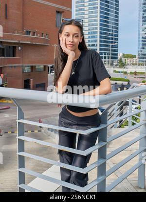 Une jeune femme aux cheveux foncés et aux lunettes sur la tête pose appuyée sur une balustrade, une main sur son visage. Elle porte un T-shirt noir et un pantalon sombre. Banque D'Images