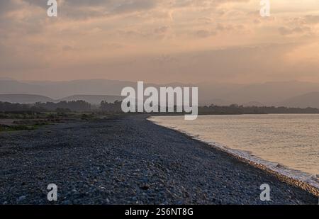 Des vagues douces tapent doucement contre la plage de galets alors que le crépuscule descend, peignant l'horizon dans des teintes chaudes. Banque D'Images