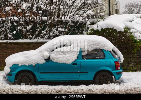 Une voiture à hayon de couleur sarcelle est garée sur une rue enneigée, lourdement chargée de neige sur son toit et son capot. La voiture est située devant une neige Banque D'Images