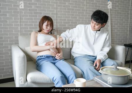 Une femme japonaise enceinte d'une vingtaine d'années portant une robe qui montre son ventre est assise sur un canapé dans un appartement, et son mari caresse son ventre. Banque D'Images