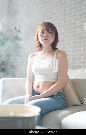 Une femme japonaise enceinte d'une vingtaine d'années et son mari, habillés pour montrer leur estomac, s'assoient sur un canapé dans un appartement. Banque D'Images