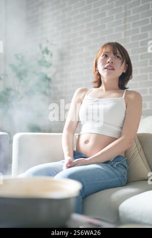 Une femme japonaise enceinte d'une vingtaine d'années et son mari, habillés pour montrer leur estomac, s'assoient sur un canapé dans un appartement. Banque D'Images