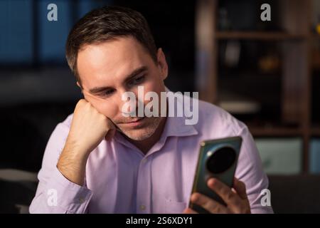 Concept Lounge. Jeune homme heureux souriant porter la chemise rose tenir dans la main utilisez le téléphone portable mobile sur fond sombre. restez à la maison plat repos détente et dépense gratuit Banque D'Images