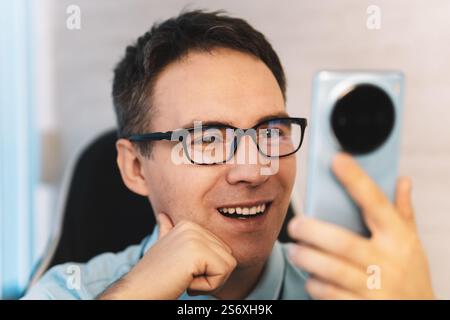 Heureux homme utilisant un smartphone à la maison, il discute messagerie en ligne sur mobile. Un jeune gars se repose dans le bureau et lit des informations d'un intelligent Banque D'Images