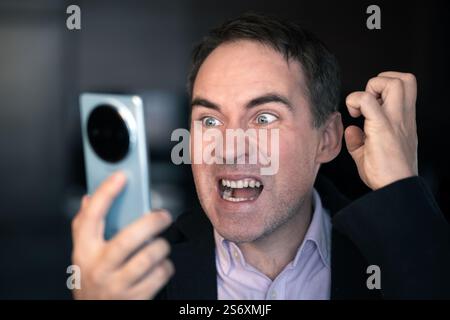 Un homme dans une chemise rose et regarde son smartphone avec un regard de choc et de fureur sur son visage. Sa bouche est ouverte, et ses yeux sont larges d'incrédulité Banque D'Images