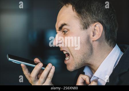 Un jeune homme avec une chemise enregistre furieusement un message vocal criant dans son smartphone. Un homme d'affaires maléfique avec un téléphone dans les mains sur un dos sombre Banque D'Images