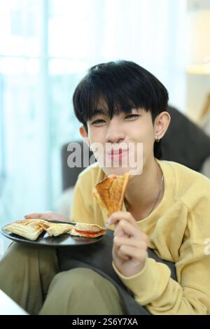 Mignon jeune homme gay appréciant des sandwichs faits maison dans un salon lumineux Banque D'Images