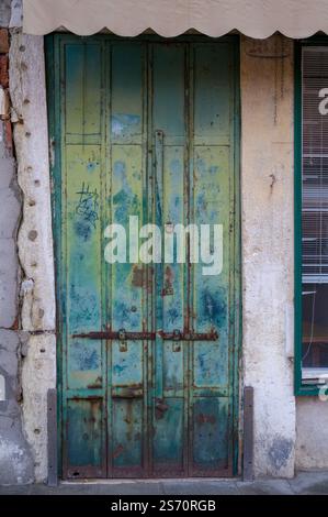 Porte en métal sarcelle usée avec quincaillerie rouillée, contre un mur de pierre en ruine et une fenêtre avec stores. Un auvent de protection surplombe l'entrée. Banque D'Images