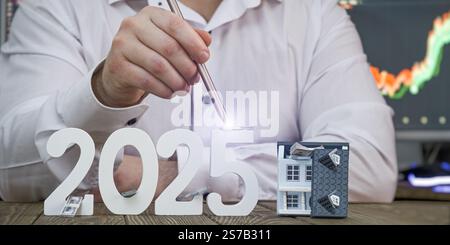 2025 immobilier et concept de croissance financière : main avec Stylus Lighting Numbers et Model House avec le graphique du marché boursier dans le Banque D'Images