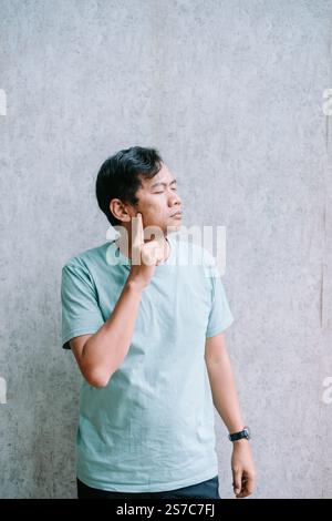 Un homme dans une chemise bleu clair se tient contre un mur texturé, pointant sa joue avec une expression réfléchie, transmettant un moment de contemplati profond Banque D'Images