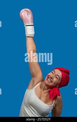 Heureuse femme âgée portant un gant de boxe rose levant le bras en célébration de la victoire sur fond bleu Banque D'Images