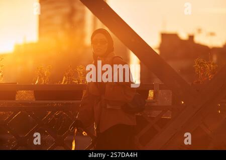 Une hijab femme dans des lunettes de soleil élégantes et une élégante tenue française, marchant dans la ville au coucher du soleil, portant un bouquet, du pain, et un journal Banque D'Images