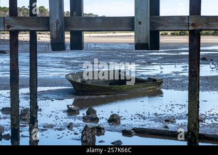 Vieux bateau repose dans l'eau peu profonde sous la jetée en bois à marée basse pendant la journée ensoleillée Banque D'Images
