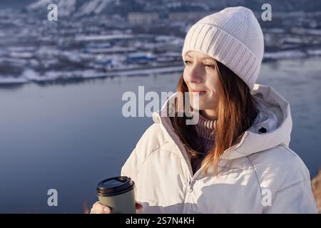 Un moment serein capturé avec une femme tenant un café chaud, admirant profondément une belle vue hivernale devant vous. Une femme d'hiver dans une veste blanche aime le Banque D'Images