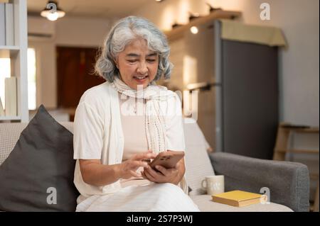 Une femme âgée asiatique heureuse et détendue dans des vêtements confortables utilise son smartphone pour discuter sur les médias sociaux ou faire des achats en ligne tout en étant assise sur un canapé dans le salon R Banque D'Images