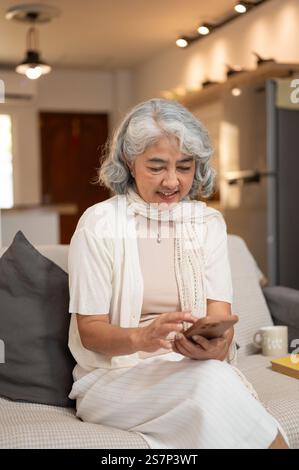 Une femme âgée asiatique heureuse et détendue dans des vêtements confortables utilise son smartphone pour discuter sur les médias sociaux ou faire des achats en ligne tout en étant assise sur un canapé dans le salon R Banque D'Images