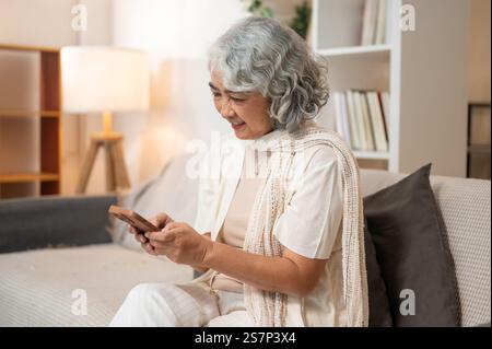 Une femme âgée asiatique heureuse et détendue dans des vêtements confortables utilise son smartphone pour discuter sur les médias sociaux ou faire des achats en ligne tout en étant assise sur un canapé dans le salon R Banque D'Images