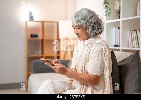 Une femme âgée asiatique heureuse et détendue dans des vêtements confortables utilise son smartphone pour discuter sur les médias sociaux ou faire des achats en ligne tout en étant assise sur un canapé dans le salon R Banque D'Images