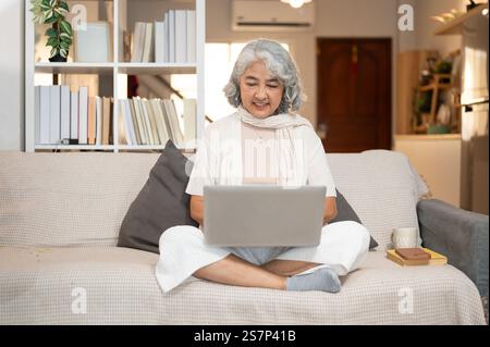 Une femme âgée asiatique heureuse utilise un ordinateur portable assis sur un canapé dans le salon. naviguer sur internet, discuter, répondre à des e-mails, travailler Banque D'Images