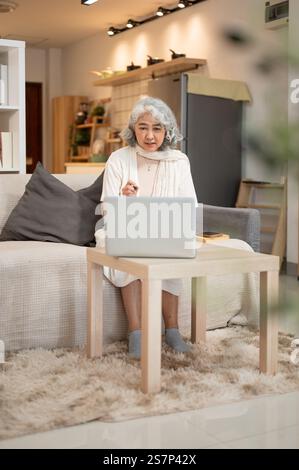 Une femme âgée asiatique heureuse utilise un ordinateur portable assis sur un canapé dans le salon. naviguer sur internet, discuter, répondre à des e-mails, travailler Banque D'Images