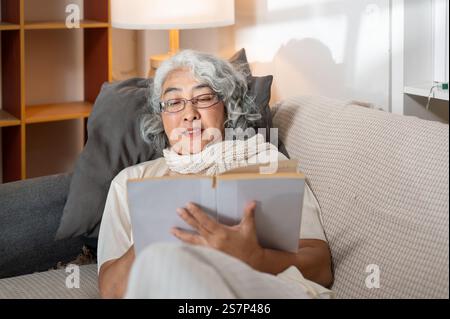 Une femme senior asiatique positive et détendue s'allonge sur un canapé dans le salon, lisant un livre la nuit. vie domestique, loisirs, retraite Banque D'Images