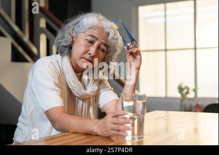 Une triste femme asiatique retraitée senior est assise sur un îlot de cuisine, regardant un verre d'eau avec un sentiment désespéré, se sentant seule après la retraite. Banque D'Images