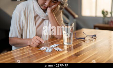 Un gros plan d'une femme asiatique âgée triste et ennuyée regarde ses médicaments sur la table avec une expression malheureuse, se sentant malade et malheureuse, et ennuyée de t Banque D'Images