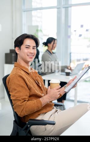 Un portrait d'un bel homme d'affaires asiatique est assis à son bureau dans le bureau avec une tasse de boisson chaude et un porte-documents dans sa main, souriant à la CA Banque D'Images
