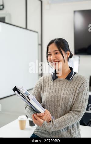 Une femme d'affaires asiatique attrayante dans un pull confortable transporte des documents et des dossiers tout en se tenant debout dans le bureau. emploi en entreprise, agence de marketing, employé Banque D'Images
