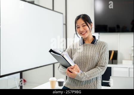 Une femme d'affaires asiatique souriante dans un pull confortable transporte des documents et des dossiers tout en se tenant debout dans le bureau. emploi en entreprise, agence de marketing, employé Banque D'Images