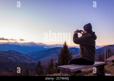 Homme prenant photo du paysage de montagne à l'aide d'un smartphone au coucher du soleil assis sur le banc profitant de la vue. Banque D'Images
