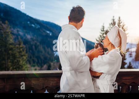 Vue arrière d'un couple tenant un chat sur le balcon de l'hôtel appréciant le paysage de montagne d'hiver portant des peignoirs. Voyagez avec votre animal de compagnie Banque D'Images
