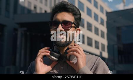 Homme indien arabe insouciant dans des lunettes de soleil porter des écouteurs écouter de la musique dans la ville ensoleillée joyeux homme d'affaires gars écoutant la chanson à l'extérieur profiter du son Banque D'Images
