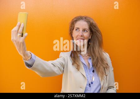 amicale femme drôle des années 60 dans la chemise bleue élégante faisant selfie sur le téléphone portable parlant sur l'appel vidéo agitant la main isolée sur fond jaune. Photo de haute qualité Banque D'Images