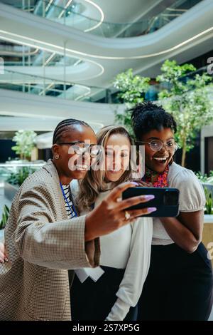 Trois collègues divers prennent un selfie dans un environnement de bureau moderne, capturant un moment de camaraderie et de travail d'équipe. Le cadre élégant avec vert Banque D'Images
