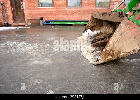 Une pelle est utilisée pour déneiger un trottoir. La pelle est grande et a beaucoup de neige dessus. La scène se déroule dans une ville, avec un buildi de briques Banque D'Images