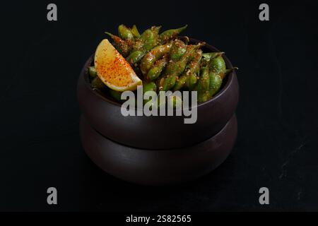 Un bol de haricots edamame assaisonnés garni d'une tranche de citron et saupoudrés de flocons de piment, créant une présentation vibrante et épicée en collation sur un Banque D'Images