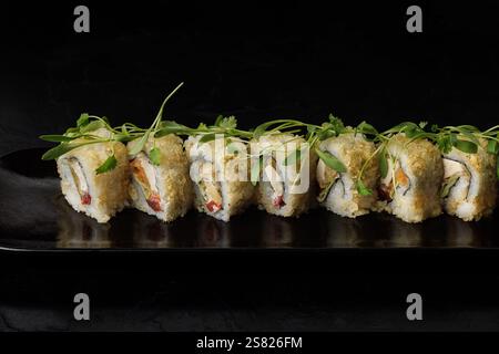Une rangée de rouleaux de sushi tempura dorés garnis de microverts frais, élégamment disposés sur une assiette rectangulaire sombre. Les petits pains sont croustillants Banque D'Images