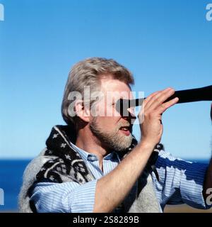 Ein Wissenschaftler beobachtet mit einem Teleskop von ihrem Aussichtspunkt auf einer grasbewachsenen Klippe aus, wie die Meereswellen auf die darunter liegenden Felsen krachen, Island um 1980. Banque D'Images