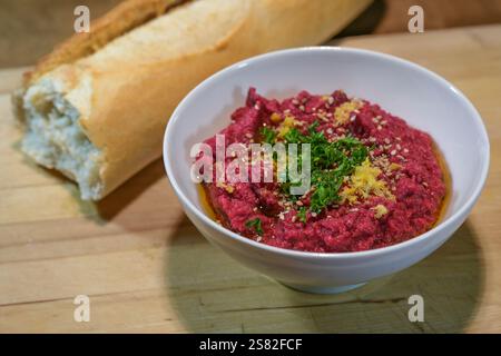 Hummus rouge fantaisie à base de pois chiches et de betteraves avec graines de sésame, garniture de persil et zeste de citron dans un bol blanc avec du pain sur une planche de bois rustique Banque D'Images