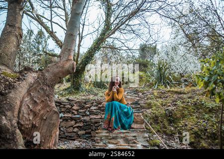 Une femme dans un haut jaune moutarde et une jupe bleue s'assoit sur un escalier en pierre entouré d'arbres, de lierre, de fleurs blanches en fleurs et d'un tronc d'arbre texturé. Banque D'Images