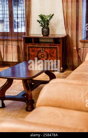 Cadre intérieur avec table basse en bois poli, canapé en cuir beige, meuble vintage avec incrustations et rideaux bruns transparents. Banque D'Images