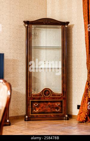 Une vitrine en bois ornée de portes vitrées et de sculptures complexes se dresse contre un mur beige à motifs, encadré par un rideau rouge et or. Banque D'Images
