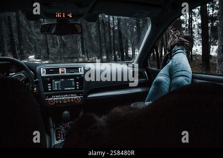 Intérieur d'une voiture avec un tableau de bord numérique, des commandes éclairées et une personne inclinée avec les pieds sur le tableau de bord. Forêt visible à l'extérieur. Banque D'Images