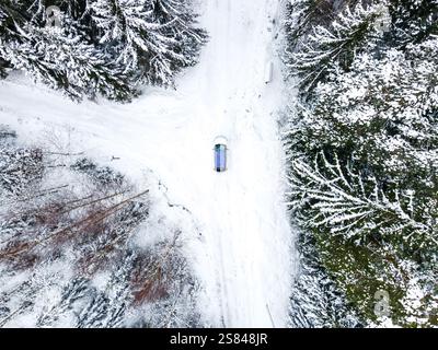 Une perspective aérienne d'une forêt enneigée entrecoupée par une route en forme de T, avec une seule voiture bleue à la jonction au milieu d'arbres chargés de neige. Banque D'Images