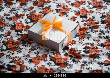 Un coffret cadeau blanc avec un ruban orange et un logo Fat Cat circulaire repose sur un tissu floral orné de roses rouges et noires sur un fond blanc. Banque D'Images